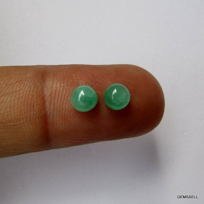 Op de afbeelding: Twee ronde, groene edelstenen, waarschijnlijk smaragden, worden op een vinger getoond. De edelstenen zijn glad en gepolijst.