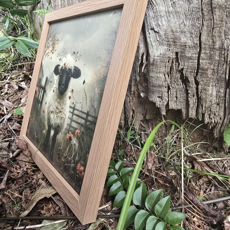 May include: A framed painting depicting a white sheep with black face and hooves, adorned with a crown of flowers. The sheep is standing in a field with a wooden fence in the background. The painting has a vintage, rustic aesthetic.