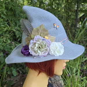 Puede incluir: Sombrero de bruja azul claro con ala ancha, decorado con detalles florales. El sombrero presenta una pluma, una mariposa y flores de tela en morado, blanco y verde. Un accesorio elegante para eventos temáticos o disfraces.