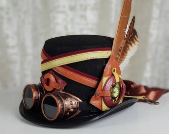 May include: Black steampunk top hat with copper-colored goggles and leather accents. The hat features a brown and yellow band, a pocket watch, and decorative feathers. A unique accessory for cosplay or themed events.