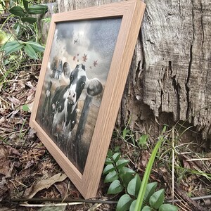 May include: A framed painting of a black and white cow standing in a field with autumn leaves in the air. The cow is looking directly at the viewer.