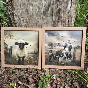 May include: Two framed canvas prints with a rustic theme. The left print features a white sheep with black face and legs standing in a field with red flowers. The right print features a black and white cow standing in a field with red flowers and a small wooden barn in the background.