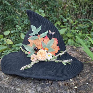 May include: A black pointed hat featuring a decorative arrangement of artificial orange and green maple leaves, a small peach-coloured flower, a white flower, a tiny orange mushroom, a brown acorn, and a small butterfly accent on the side.