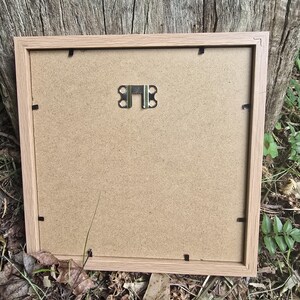 May include: A light brown wooden picture frame with a brown cardboard backing. The frame has a metal hanging hook on the back.