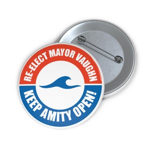 Può includere: Un bottone rotondo con un design rosso e blu. Il bottone recita "RE-ELECT MAYOR VAUGHN" in rosso e "KEEP AMITY OPEN!" in blu. Una grafica a onda blu è al centro. Un secondo bottone è visibile sullo sfondo.