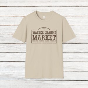 Puede incluir: Una camiseta beige de manga corta con un estampado gráfico marrón oscuro en el pecho. El diseño es un letrero rectangular que dice Walter Chang's Market, Est. 1984, General Store, Video Rental, Cold Beer, Perfection, NV y Watch Out For The Graboids.