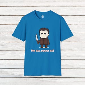 Puede incluir: Camiseta azul con una ilustración de dibujos animados de un personaje que se asemeja a Michael Myers, sosteniendo un cuchillo. El personaje es marrón con una máscara blanca. El texto "FUN SIZE, DEADLY SIZE" está impreso en rosa.