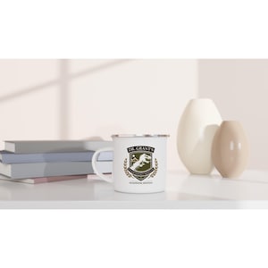 Pode incluir: Caneca de esmalte branco com borda e alça prateadas. A caneca apresenta um brasão com um gráfico de dinossauro, o texto "DR. GRANT'S PALEONTOLOGY INSTITUTE" e "SNAKEWATER, MONTANA". A caneca está em uma superfície branca.