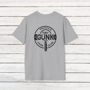 May include: A gray t-shirt with a black circle design on the back. The circle says "Private Eye Tony Gunka Staten Island" and has a magnifying glass icon in the center.