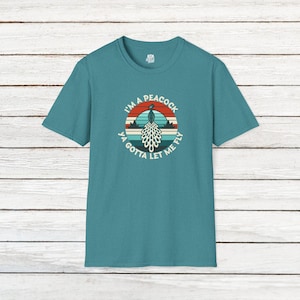 May include: A teal blue t-shirt featuring a vintage-style graphic of a peacock with the text "I'm a peacock ya gotta let me fly".