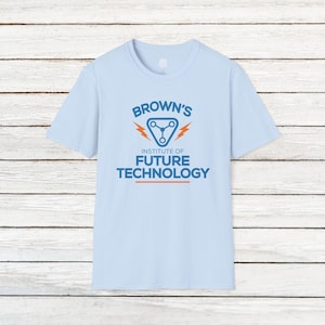 May include: Light blue t-shirt with the text "BROWN'S INSTITUTE OF FUTURE TECHNOLOGY" in blue and orange. The design includes a stylized triangle with a network symbol and lightning bolts. The shirt is displayed on a light wood background.