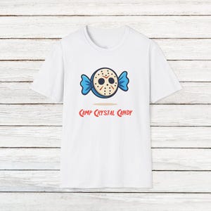 Puede incluir: Camiseta blanca con un diseño de dibujos animados de un caramelo con forma de máscara de hockey, envoltorio azul y el texto "Camp Crystal Candy" en rojo. El diseño está centrado en la parte delantera de la camiseta.