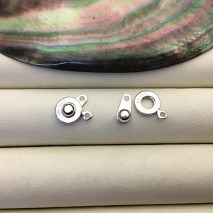 Puede incluir: Dos cierres de metal plateados para la elaboraci&oacute;n de joyas. Un cierre tiene una forma redonda con una peque&ntilde;a bola en el centro y el texto "PAT. 2001" grabado en el lateral. El otro cierre tiene una forma de anillo con una peque&ntilde;a bola en el lateral.