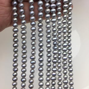 Puede incluir: Múltiples hilos de perlas de agua dulce de color gris claro y brillante dispuestos en filas verticales. Cada hilo consta de numerosas perlas pequeñas, redondas y ligeramente irregulares con una superficie lisa y reflectante de tono gris plateado.