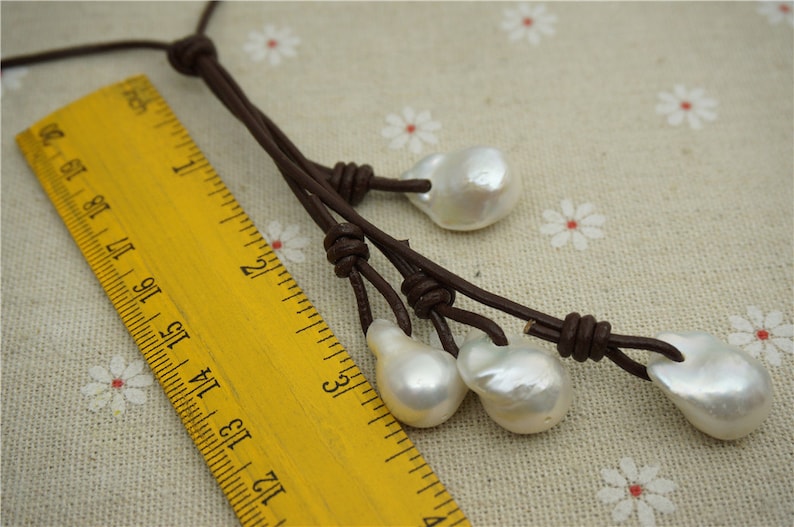 Puede incluir: Un collar de cuero marr&oacute;n con cuatro perlas barrocas blancas que cuelgan de &eacute;l. El collar est&aacute; atado con nudos y se mide contra una regla amarilla.