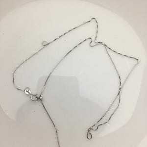 Può includere: Una collana color argento con un delicato design a catena. La collana presenta una piccola chiusura e una lunghezza regolabile. La catena è su una superficie bianca.