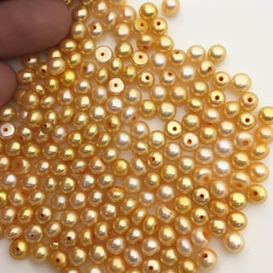 Może przedstawiać: Stosik złotych pereł, każda z gładką, perłową powierzchnią. Perły są rozrzucone na białym tle.