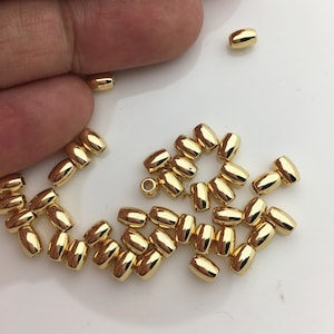 Puede incluir: Un grupo de cuentas de metal de color dorado, algunas con un agujero en el centro, están esparcidas sobre una superficie blanca. Las cuentas son de forma ovalada y tienen un acabado liso y brillante.