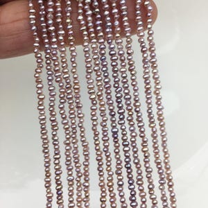 May include: Strands of small, iridescent, pinkish-purple pearls. The pearls are strung together, creating multiple vertical lines. The pearls have a slightly irregular shape and a subtle luster. The background is a plain white surface.