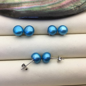 May include: Several pairs of blue pearl stud earrings. The earrings have a lustrous, iridescent finish. The posts and backs are silver-colored. The earrings are displayed on a white surface.