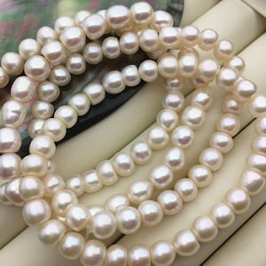 Puede incluir: Una hebra de perlas de agua dulce blancas, con una forma redonda y lisa. Las perlas están dispuestas en un patrón suelto y fluido, creando un aspecto delicado y elegante.