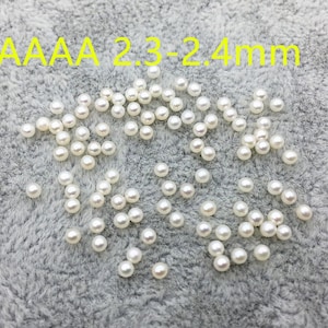 May include: A scattering of white round pearls on a gray fuzzy surface. The pearls are approximately 2.3-2.4 mm in size. The text "AAAA 2.3-2.4mm" is visible in the image.