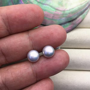 Op de afbeelding: Twee ronde, lichtgekleurde parel oorbellen. De parels hebben een subtiele iriserende glans, die hints van blauw en paars reflecteert. De oorbellen worden in een hand gehouden, met een vage achtergrond van een schelp.
