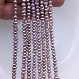 Peut inclure: Plusieurs brins de perles d'eau douce rose-lavande. Les perles de forme ovale ont un éclat brillant. Les brins sont tenus dans une main, montrant la couleur et la forme des perles. Les perles sont enfilées, prêtes pour la fabrication de bijoux.