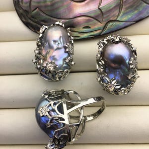 May include: Three silver rings with iridescent gray pearls set in a floral design. The rings are on a white surface.