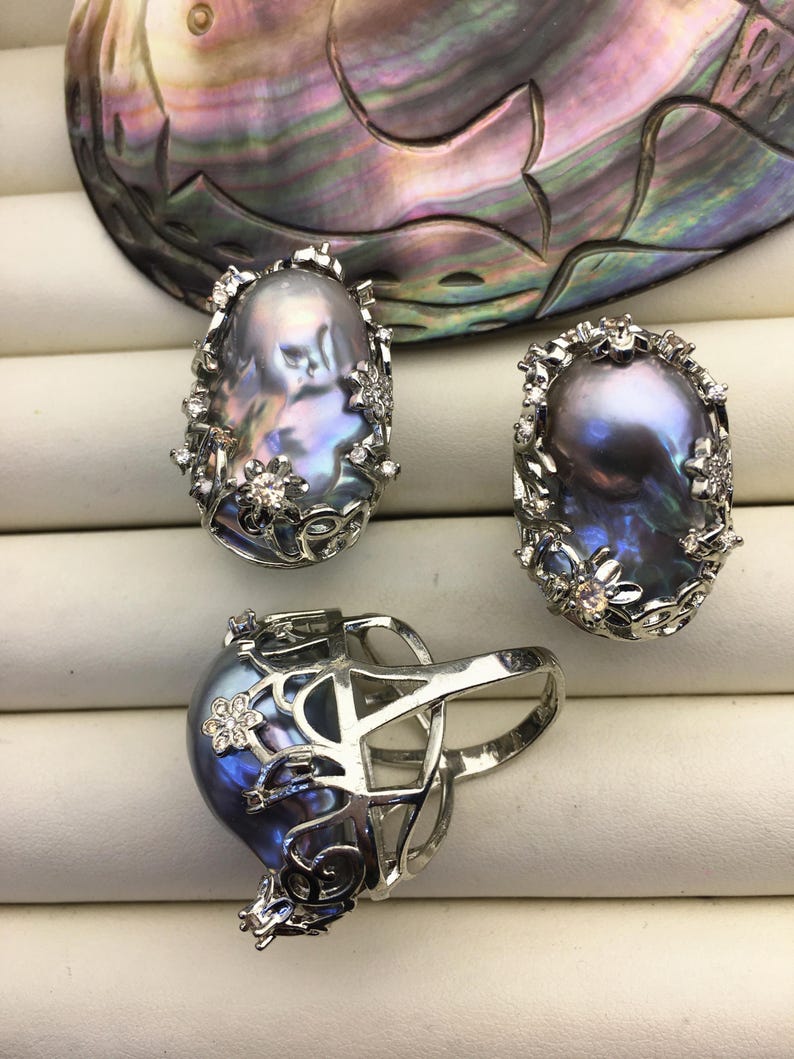 May include: Three silver rings with iridescent gray pearls set in a floral design. The rings are on a white surface.