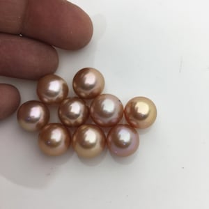 May include: A group of nine round, iridescent pearls in shades of pink and peach. The pearls are smooth and glossy, and they are arranged in a cluster on a white surface.