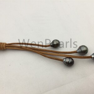 May include: A brown leather cord necklace with five dark gray pearls hanging from it. The cord is knotted at the top and the pearls are evenly spaced.