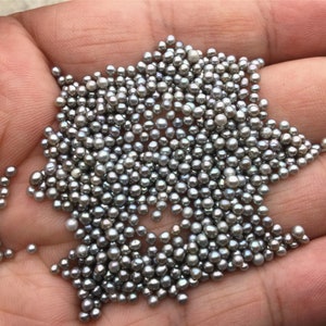 Puede incluir: Un puñado de pequeñas cuentas de color gris plateado. Las cuentas son redondas y parecen estar hechas de un material metálico. Están agrupadas, con algunas que se caen de la mano. Las cuentas son de tamaño y forma uniformes, creando una superficie texturizada.