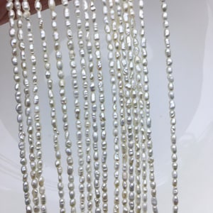 Puede incluir: Hilos de perlas blancas de forma irregular. Las perlas están ensartadas, creando múltiples líneas verticales. Las perlas tienen un brillo natural y varían en tamaño y forma, con una mezcla de formas ovaladas y oblongas. El fondo es una superficie blanca lisa.