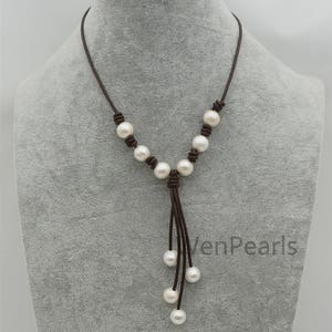 May include: A brown leather necklace with white pearls. The necklace has a tassel with five pearls at the end. The necklace is on a mannequin.
