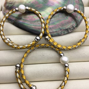 May include: Three braided leather bracelets with silver accents and white pearl accents. The bracelets are on a white surface.