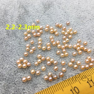 Puede incluir: Primer plano de un grupo de pequeñas perlas redondas de color rosa pálido, que miden 2,2-2,3 mm, esparcidas sobre una superficie gris texturizada. Una cinta métrica amarilla es visible en la esquina inferior derecha.