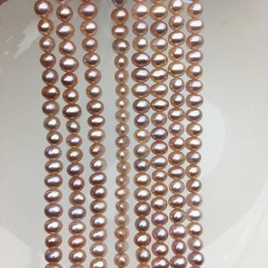 May include: Several strands of pink freshwater pearls. The pearls are oval-shaped and have a lustrous sheen. The strands are hanging against a white background, showcasing the pearls' color and shape. The pearls vary slightly in size.