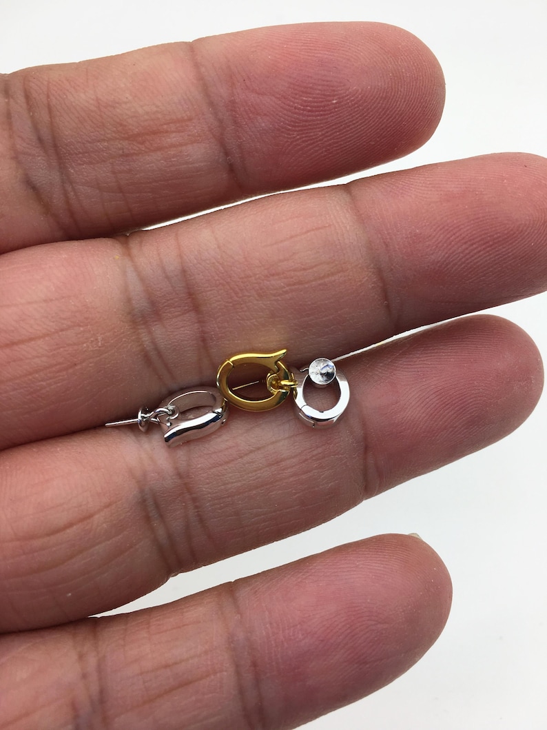 Pu&ograve; includere: Primo piano di tre piccole chiusure per gioielli in tonalit&agrave; argento e oro. Le chiusure sono in diversi stili, tra cui un moschettone, una forma a ferro di cavallo e una chiusura rotonda con una piccola pietra trasparente. Le chiusure sono tenute in una mano.