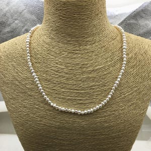 Può includere: Una delicata collana composta da piccole perle bianche e rotonde. Le perle sono infilate insieme per formare un'unica fila, esposta su un busto marrone. La collana è un classico della gioielleria.