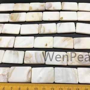 Może przedstawiać: Szereg prostokątnych koralików muszlowych w kolorze białym i kremowym. Koraliki mają około 2,5 cm długości i 1,25 cm szerokości. Koraliki są ułożone w rzędzie i oddzielone cienkim sznurkiem. Linijka jest widoczna na dole obrazu, pokazując rozmiar koralików.