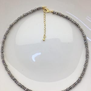 May include: A necklace composed of small, irregular, grey-toned pearls strung together in a continuous circular strand. The piece features a gold-toned metal lobster claw clasp and an adjustable extender chain with a small teardrop-shaped charm.