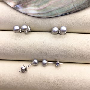 May include: Several pairs of pearl earrings are displayed. The earrings feature small, round, gray pearls set on silver posts. Some have decorative silver backings. The pearls have a subtle luster, and the earrings are arranged on a light beige surface.