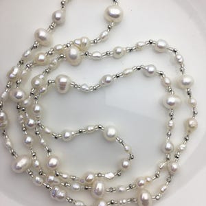 Pode incluir: Um colar de pérolas brancas com várias voltas de pérolas de tamanhos diferentes. As pérolas são enfiadas em uma corrente prateada.