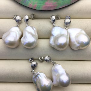 May include: Three pairs of white baroque pearl earrings with silver-toned metal accents. The pearls have an irregular shape and a lustrous surface. The earrings are displayed on a cream-colored surface.