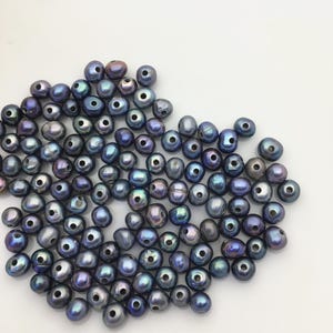 Könnte beinhalten: Eine Sammlung dunkelblauer, schillernder Süßwasserperlen. Die Perlen sind oval geformt mit natürlichem Glanz und sichtbaren Bohrlöchern, verstreut auf einer weißen Oberfläche. Diese Perlen eignen sich für die Schmuckherstellung.