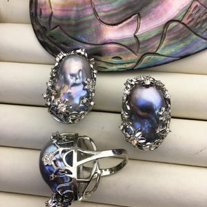 May include: Three silver rings with a large, iridescent, oval-shaped pearl in each. The rings are set with small, clear stones and have a floral design.