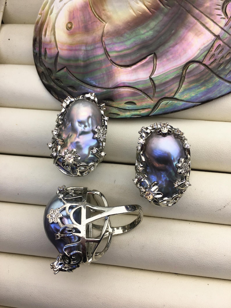 May include: Three silver rings with a large, iridescent, oval-shaped pearl in each. The rings are set with small, clear stones and have a floral design.