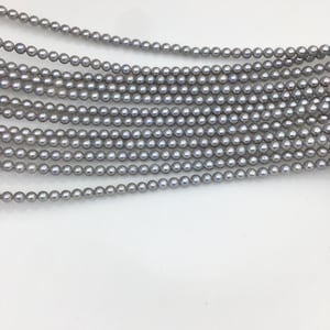 Puede incluir: Varias hebras de perlas grises, cada una con un brillo sutil, se muestran sobre un fondo blanco. Las perlas son uniformes en tamaño y forma, creando un aspecto clásico y elegante. La imagen resalta la belleza natural de las perlas.