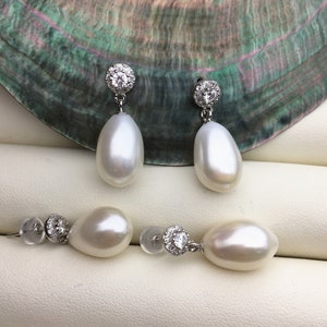 May include: A pair of white pearl drop earrings with a silver setting and a small cluster of clear cubic zirconia stones. The earrings are displayed on a white surface.